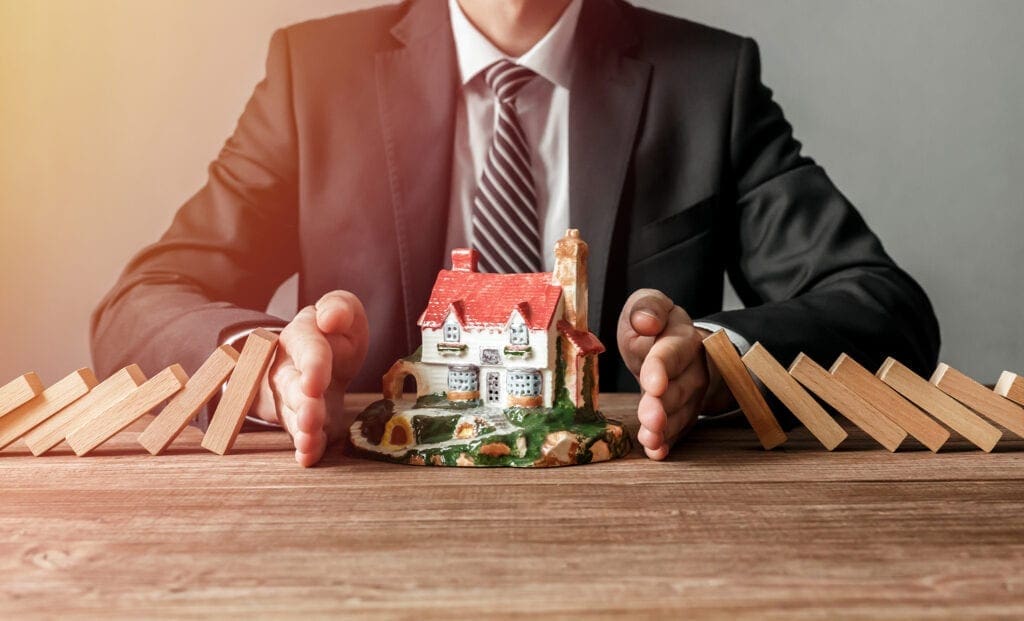 Close-up of a man stopping the wooden blocks from falling on house model. House insurance and security concept.